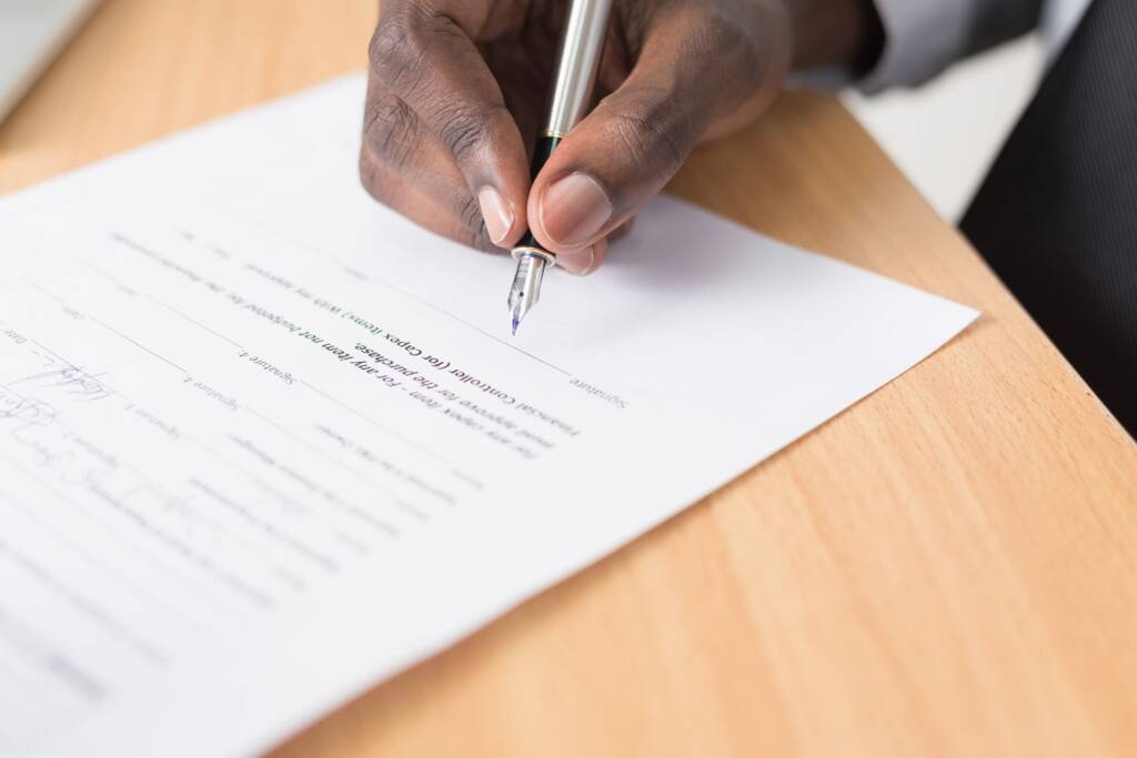 Person signing a document with a pen on a wooden table.