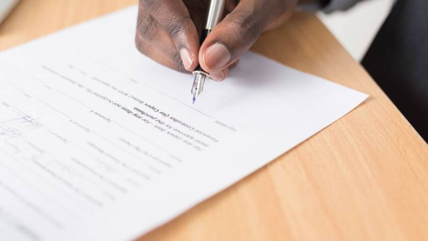 Person signing a document with a pen on a wooden table.
