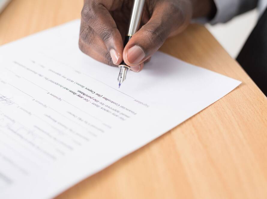 Person signing a document with a pen on a wooden table.