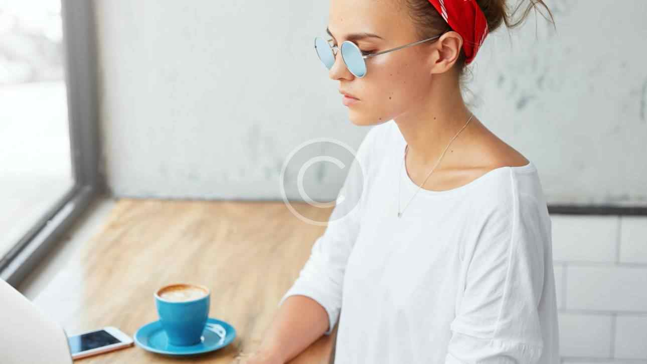 Woman in glasses and red headband working on laptop with coffee.