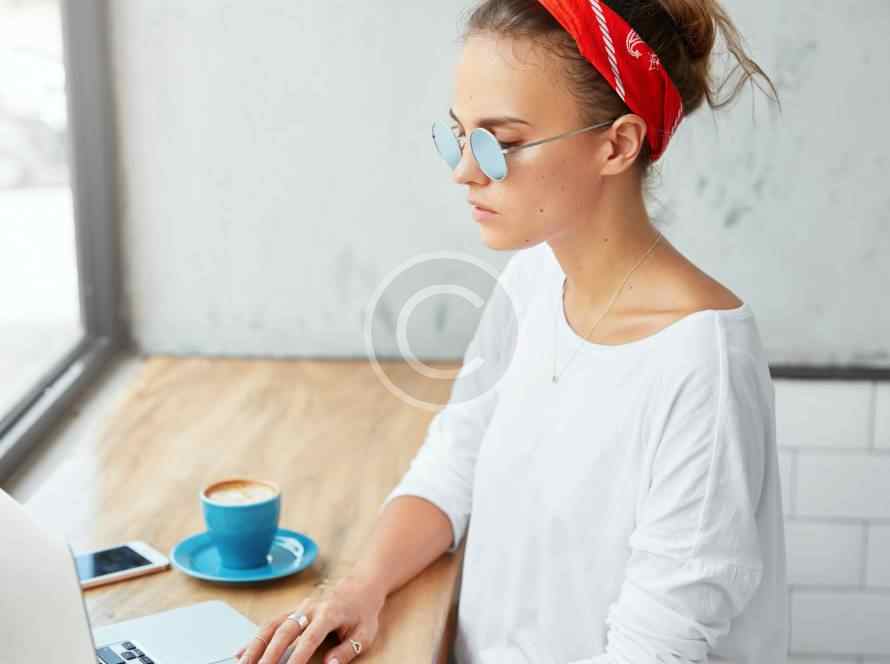 Woman in glasses and red headband working on laptop with coffee.