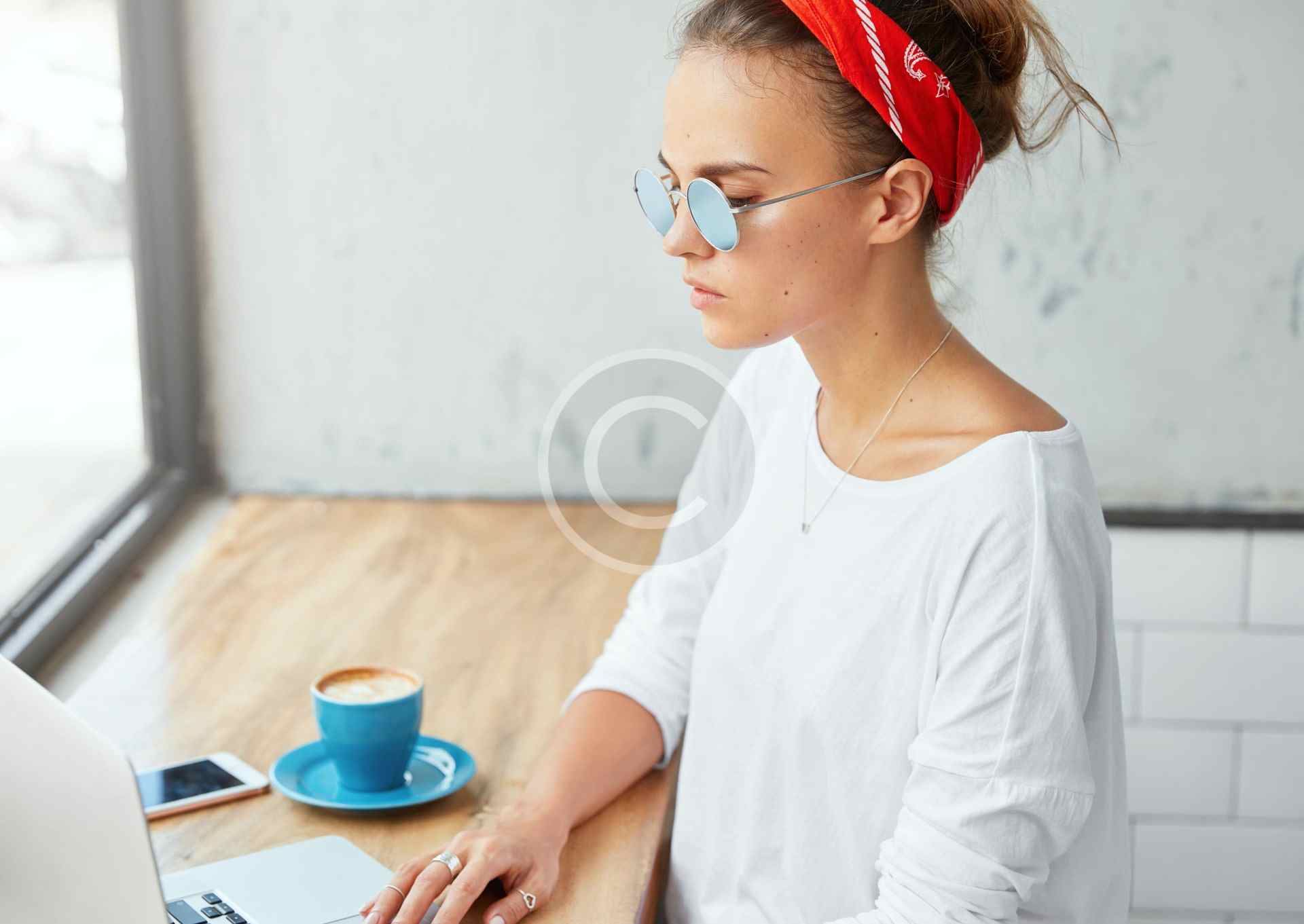 Woman in glasses and red headband working on laptop with coffee.