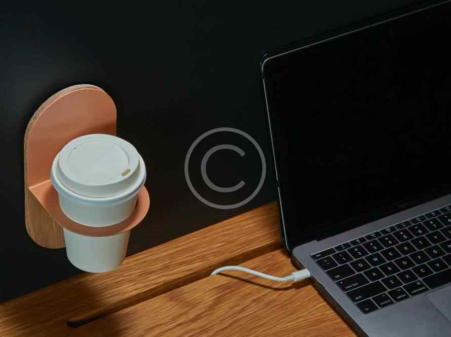 Hand holding a coffee cup near a laptop and charging cable on a dark surface.