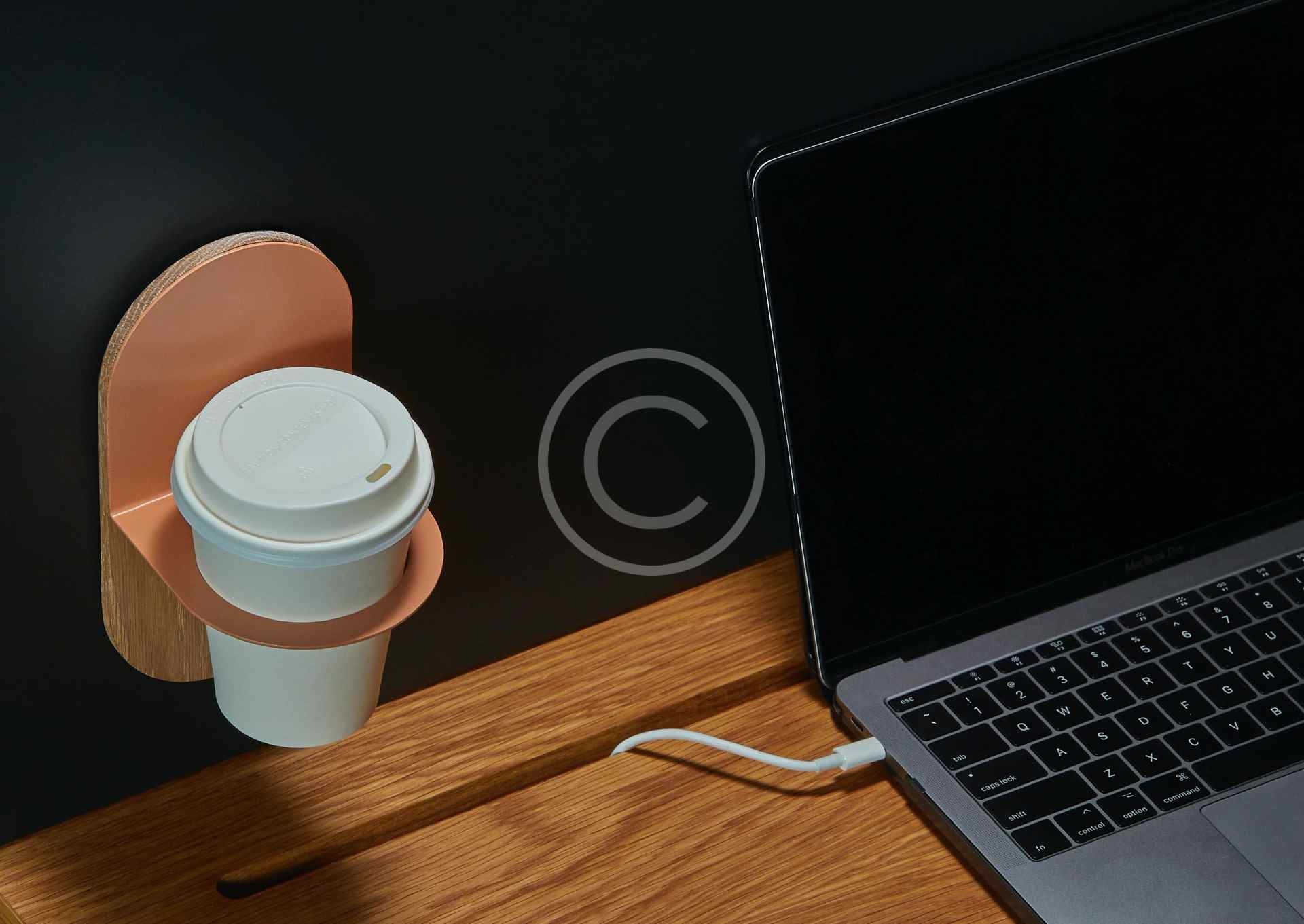 Hand holding a coffee cup near a laptop and charging cable on a dark surface.