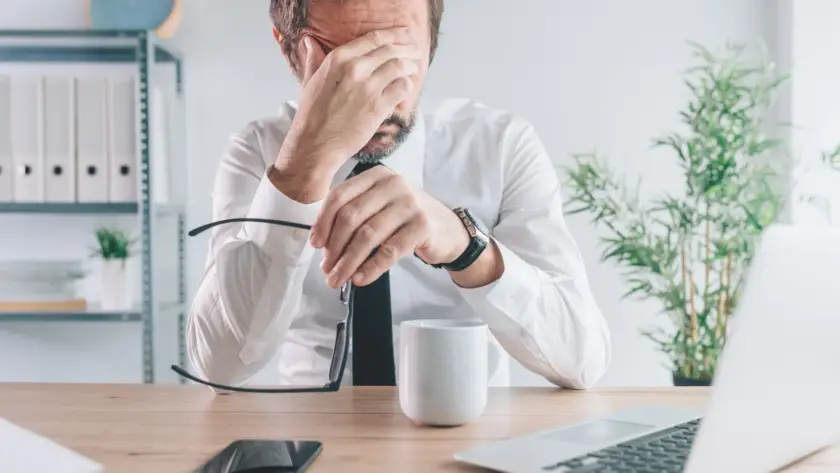 Stressed man sitting at desk with hand on face, surrounded by work items.
