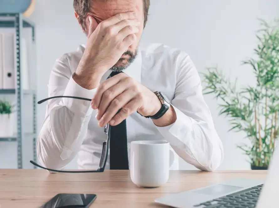 Stressed man sitting at desk with hand on face, surrounded by work items.