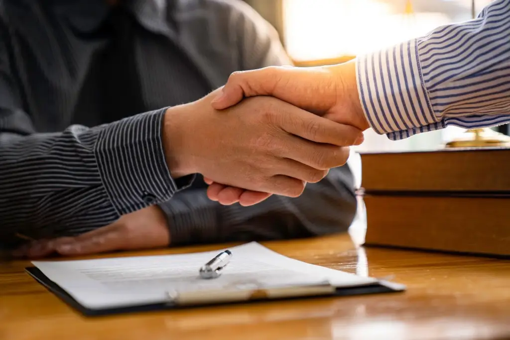 Two people shaking hands over a contract on a table.