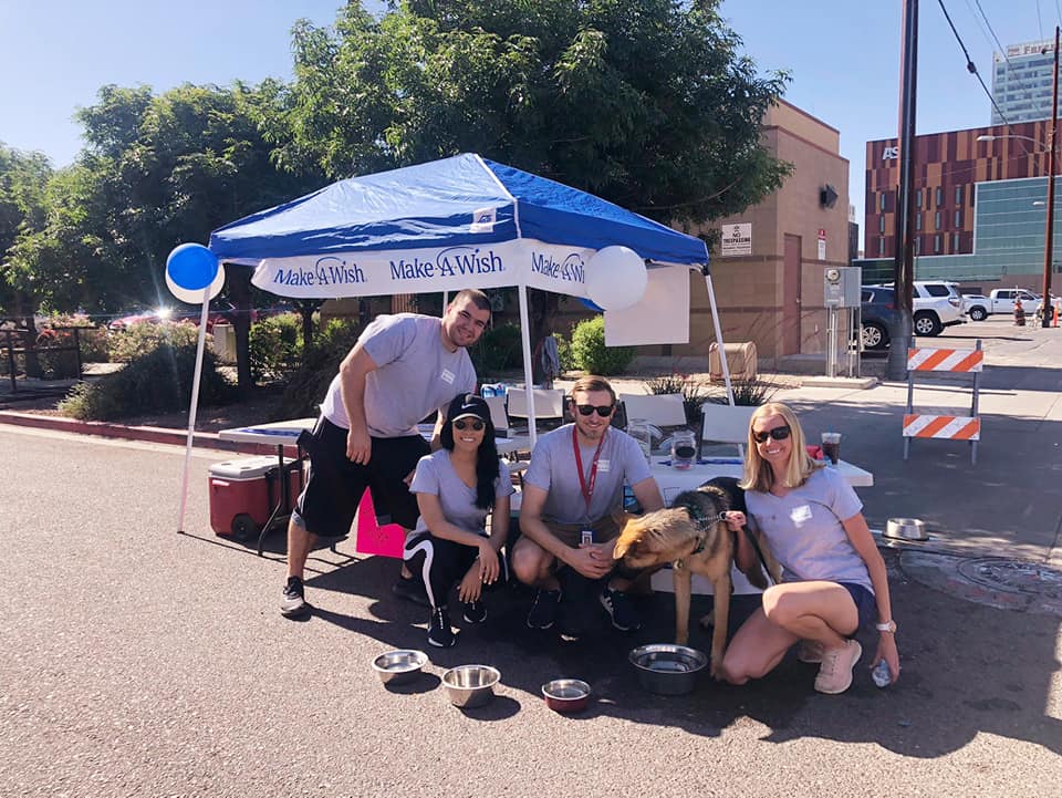 Four people sitting and standing by a tented booth outdoors on a sunny day.