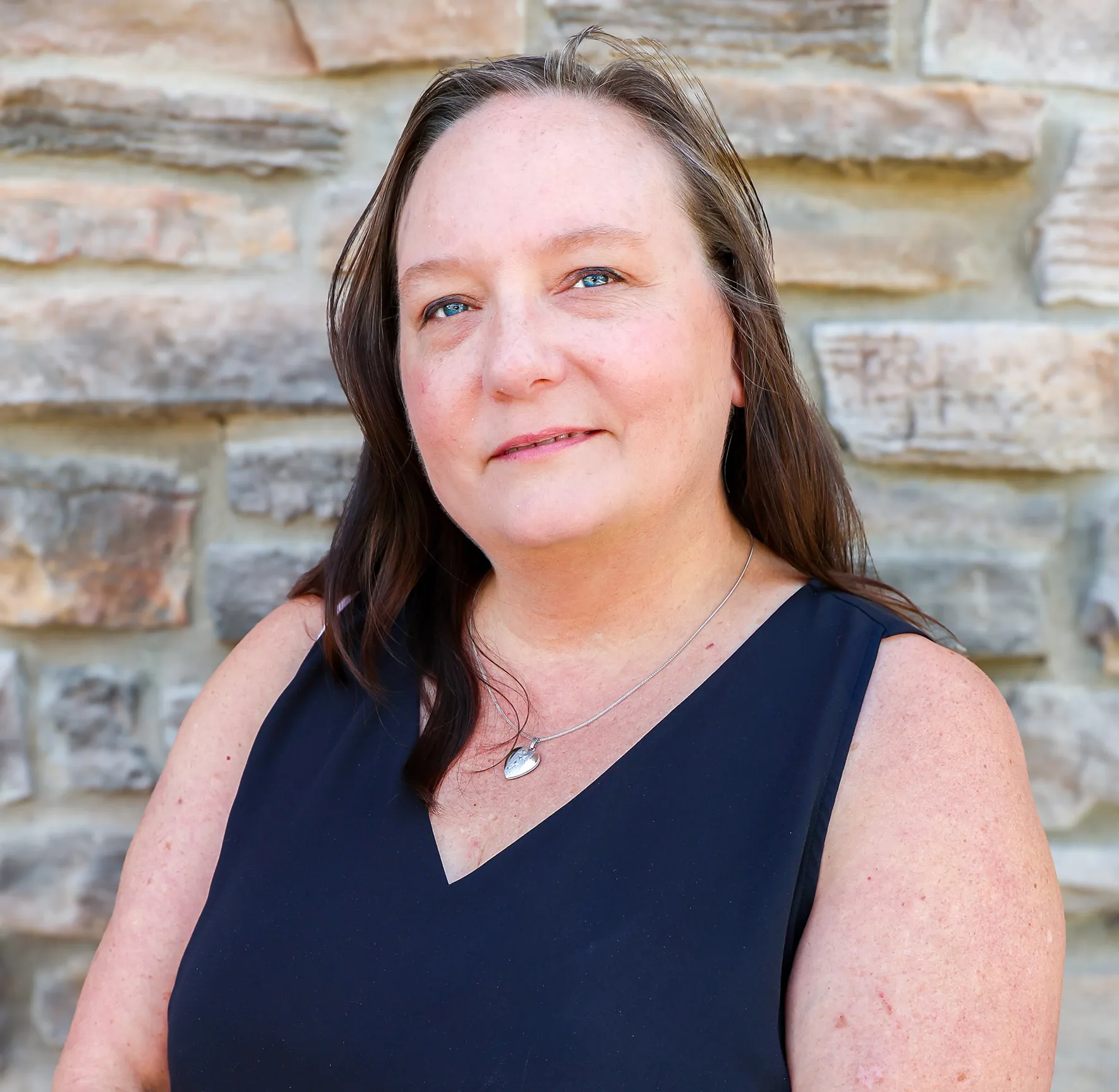 Woman with long brown hair in a black sleeveless top standing against a stone wall.