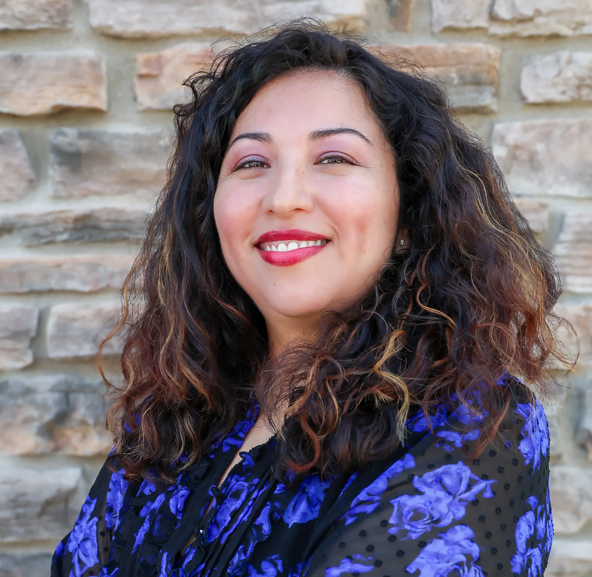 Smiling woman with curly hair and floral jacket in front of a stone wall.