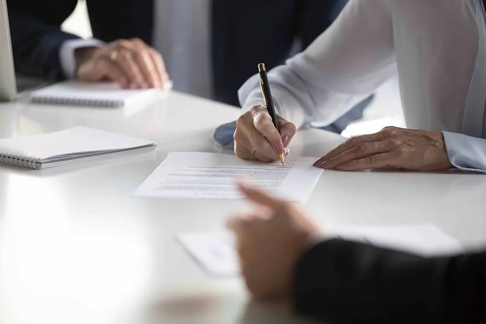Person signing a document on a desk with others present.