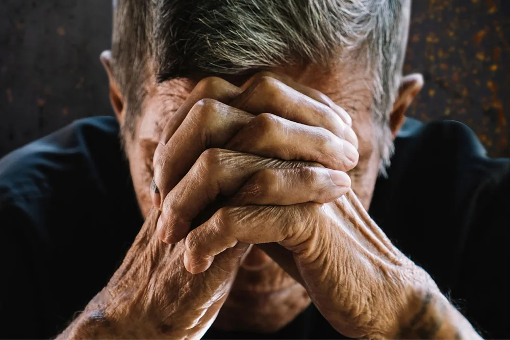 An elderly person with hands clasped over their face in contemplation.