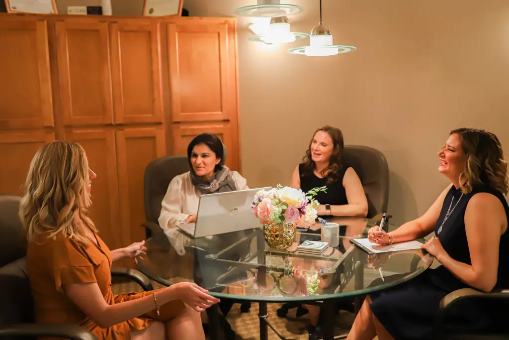 Four women chat around a glass dining table in a cozy, warmly lit kitchen.