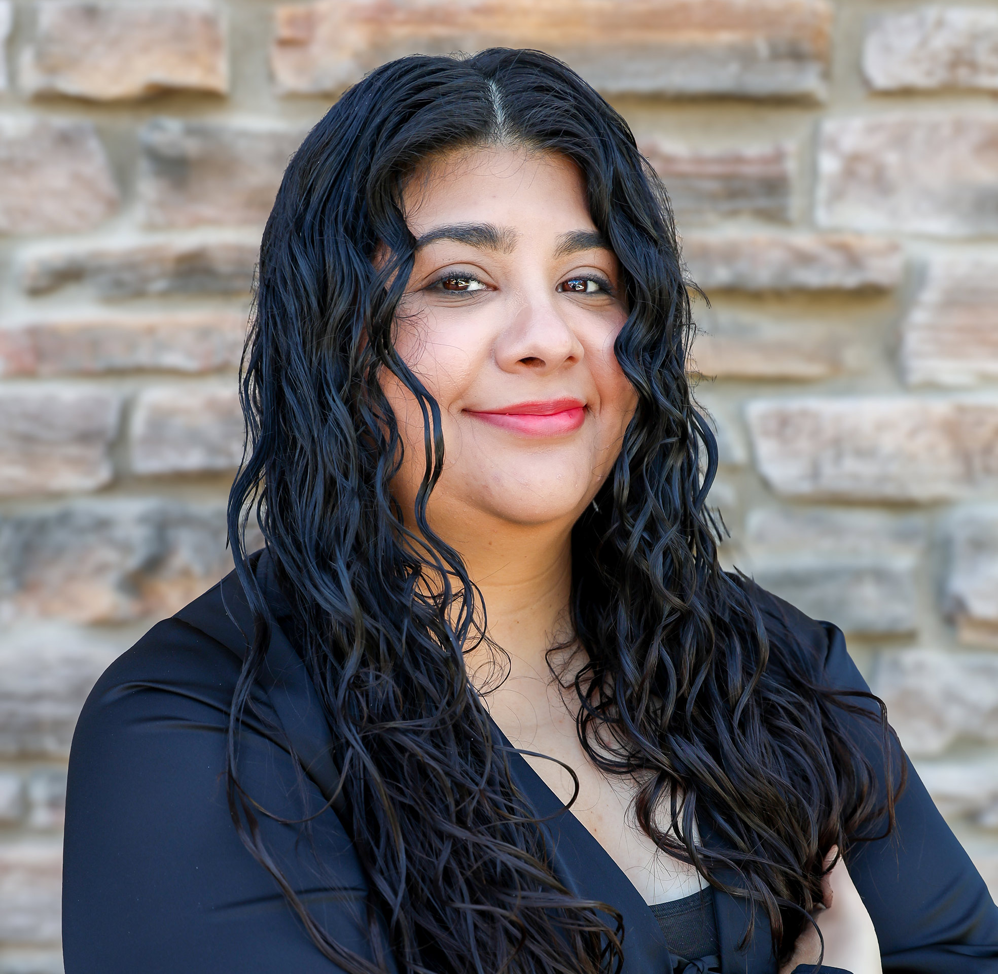 Confident woman with long curly hair stands smiling against a stone wall.